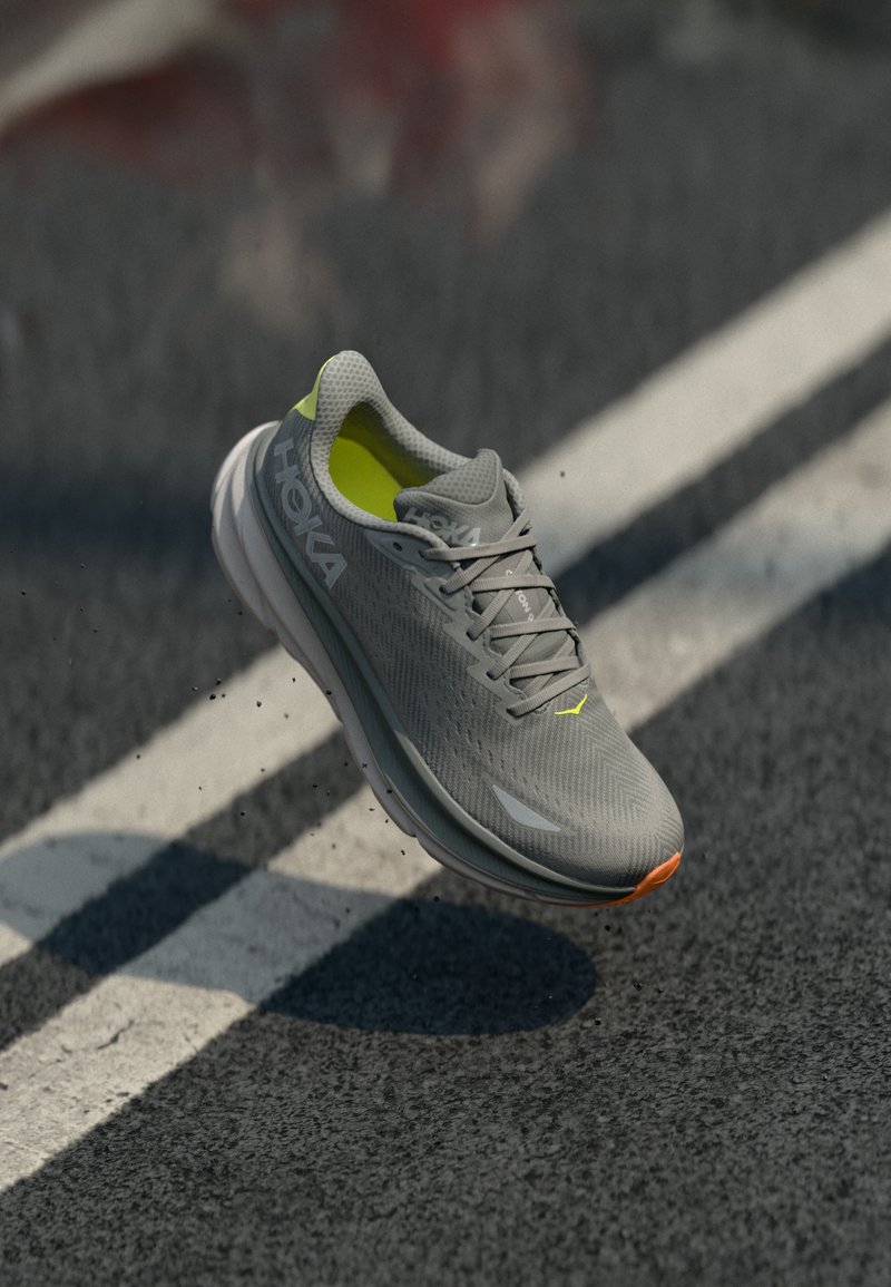 Scarpa atletica grigia con una tomaia in rete traspirante, accenti in neon giallo, suola ammortizzata e dettagli texturizzati, mostrata sospesa sopra il pavimento.