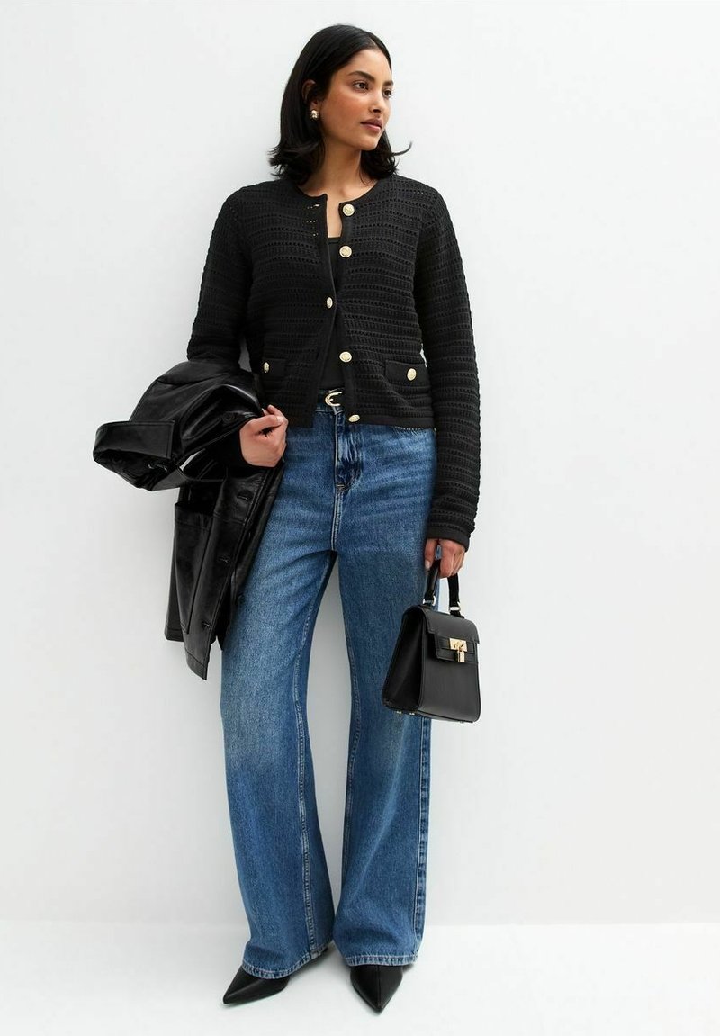 Gilet noir texturé avec des boutons dorés, associé à un jean bleu évasé taille haute. Veste en cuir noire drapée sur le bras et petit sac.
