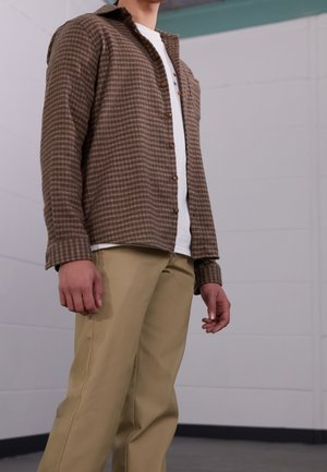 Mężczyzna w beżowych spodniach, białej koszulce i brązowej koszuli w kratę stoi w pomieszczeniu na tle jednolitej ściany.