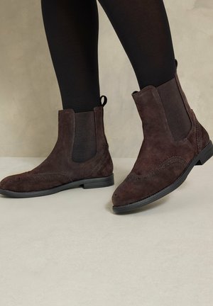 Bottines en daim brun foncé avec panneaux latéraux élastiques et détails brogue, portées avec des collants noirs sur un sol clair.