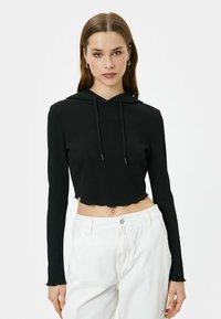 Sudadera corta negra con textura acanalada, mangas largas y un dobladillo ondulado. Presenta una capucha con cordón y un acabado de tela suave.