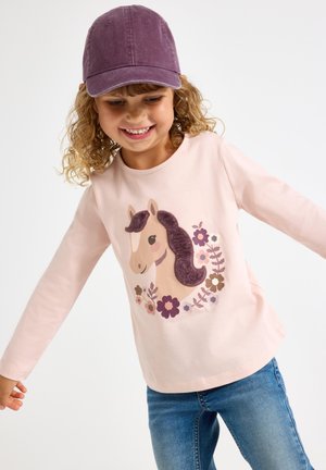 Ung pige smiler og balancerer med armene ud, iført en lilla kasket, lyserød skjorte med hest og blomsterdesign samt blå jeans.