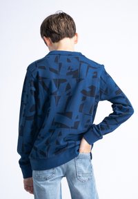Sweatshirt navy con motivi astratti neri, colletto rotondo, maniche lunghe e polsini a costine; abbinato a jeans azzurri chiari.