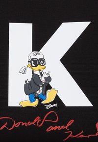 Un fond en tissu noir présente une grande lettre blanche "K." Une canard de dessin animé portant des lunettes de soleil, un manteau noir et des bottes bleues, tient un chat.