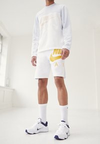 Bílá dlouhá košile Nike s síťovými detaily, žluté logo; odpovídající bílé šortky s velkým žlutým logem. Bílé ponožky a tenisky.