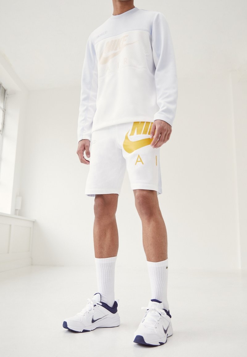 Bílá dlouhá košile Nike s síťovými detaily, žluté logo; odpovídající bílé šortky s velkým žlutým logem. Bílé ponožky a tenisky.