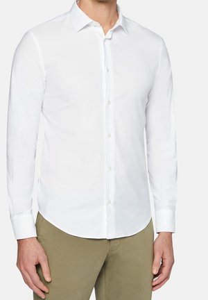 Mann trägt ein eng anliegendes, weißes Hemd mit langen Ärmeln und Knöpfen sowie olivgrüne Hose, steht mit entspannten Armen an den Seiten.