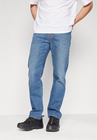 Hombre vistiendo jeans de color azul claro, zapatillas negras y una camiseta blanca, de pie con las manos detrás de la espalda contra un fondo liso.