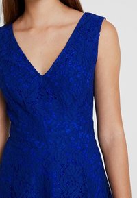 Niebieska sukienka koronkowa z dekoltem w serek i bez rękawów, prezentująca misternie zdobione wzory kwiatowe oraz dopasowany krój.