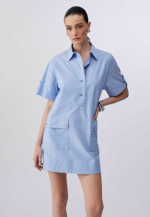 Kobieta o ciemnych włosach w jasnoniebieskiej sukience z krótkim rękawem, dużymi przednimi kieszeniami i srebrnymi guzikami, stojąca na tle jednolitego białego tła.