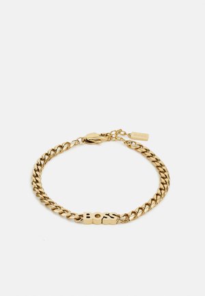 Bracciale in metallo dorato con design a maglie piatte e un ciondolo centrale che scrive "BOSS". Include una chiusura a fibbia e un'etichetta con il logo.
