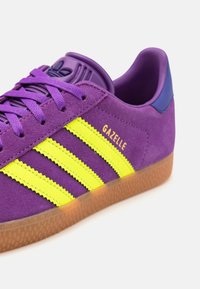 Lila velúr sneaker sárga három csíkkal, gumitalppal és kék akcentussal a saroknál. A "GAZELLE" logó aranyszínűen megvarrva.