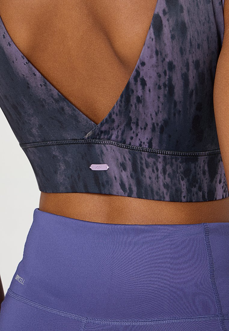 Reggiseno sportivo nero e viola a fantasia con schiena a V profonda, caratterizzato da cuciture a contrasto e un piccolo etichetta con logo all'orlo. Abbinato a leggings viola.