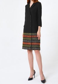 Svart skräddarsydd klänning med tre knappar, trekvartslånga ärmar och en vävd flerfärgad nederdel som har ränder i orange, grönt och grått.