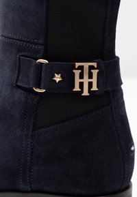 Námořnické semišové boty s černým textilním panelem, zlatým sponou s iniciály, hvězdným akcentem a viditelnými švy.