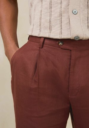 Bărbat îmbrăcat în pantaloni de culoare ruginiu cu buzunare laterale și o cămașă tricotată bej cu mâneci scurte, dungi verticale și nasturi.