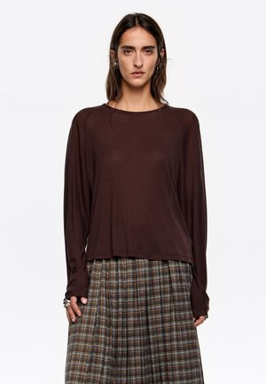 Brązowa bluzka z długim rękawem o luźnym fasonie i okrągłym dekolcie, zestawiona z plisowaną spódnicą w kratę z ziemistymi tonami i naprzemiennymi paskami.
