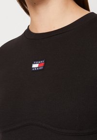 Black ribbed knit top featuring a round neckline and a small embroidered logo patch in red, white, and blue on the chest area.