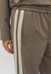 Bruna träningsbyxor med en vävd beige rand och grå accenter. Resårmidja med dragsko och en sidoficka.