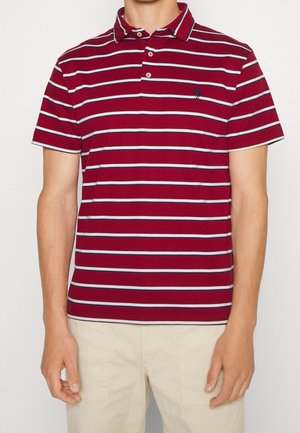Man draagt een rood-wit-marineblauw gestreept poloshirt met korte mouwen en een beige broek tegen een effen achtergrond.