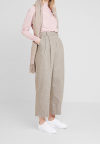 Pantaloni beige a vita alta con pieghe, dal taglio sartoriale, abbinati a una maglietta rosa chiaro a maniche lunghe e a una sciarpa beige, completati da sneaker bianche.