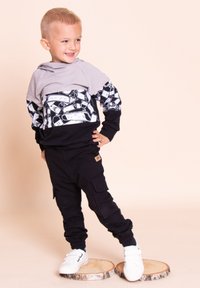 Szara bluza z kapturem z czarno-białym nadrukiem graficznym, w połączeniu z czarnymi spodniami typu cargo. Buty są białe z czarnymi akcentami.