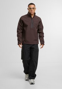 Férfi barna cipzáras dzsekiben, fekete cargo nadrágban és fehér sportcipőben áll egy sima világosszürke háttér előtt.