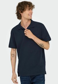 Navy short-sleeve polo shirt with a textured fabric, collar, and a chest pocket. Model is wearing denim jeans and displaying tattoos on arms.