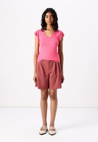 Top rosa de canalé con escote en V y mangas con volantes, combinado con pantalones cortos marrones holgados. La modelo está de pie frente a un fondo neutro, vistiendo zapatos planos.