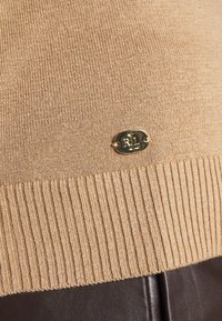 Pull en tricot beige avec ourlet côtelé et petite étiquette ovale dorée de la marque embossée "RL" en bas devant.