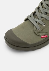 Scarpa di tela verde con punta in gomma, suola textured e occhielli intrecciati per i lacci. Presenta un piccolo logo rosso vicino al lato.