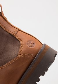 Bottines en cuir marron avec des panneaux latéraux en textile marron, des coutures contrastantes et un logo de marque sur le côté extérieur. Semelle en caoutchouc.
