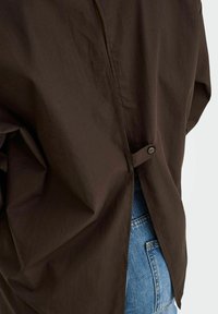 Hnědá košile na zapínání s uvolněným střihem, obsahující výřez vzadu a detail s knoflíkem. Texturovaná látka, spárována s modrými džínami.