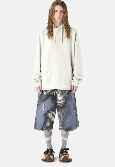 Jeune personne aux cheveux longs et bouclés portant un sweat à capuche oversize écru, un short large à motifs noir et blanc, des chaussettes grises et des baskets noires.