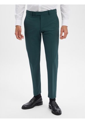 Homme portant un pantalon slim vert foncé, des chaussures habillées noires et une chemise blanche, debout devant un fond blanc uni.