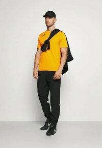 Cămașă scurtă, de culoare portocalie strălucitoare, pantaloni negri și șapcă neagră. Pulover negru aruncat pe umeri. Pantofi sport negri cu accente gri.