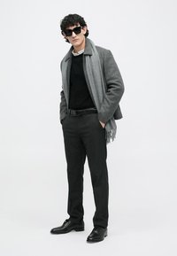 Casaco cinzento com um suéter escuro, calças escuras e sapatos pretos, complementado por um cachecol cinzento. Os acessórios incluem óculos de sol pretos.