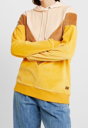 Person with arms crossed wearing a yellow, brown, and beige color-blocked hoodie paired with loose blue jeans.