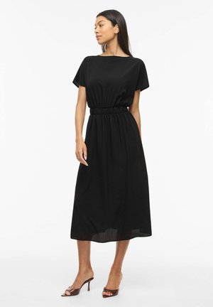 Kvinne iført en svart midi-kjole med korte ermer og samlet midje, stående og ser til siden, kombinert med brune åpne hæler.