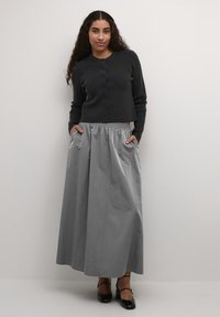 Zwarte geribbelde cardigan met knopen, gecombineerd met een lange, grijze, geplooide rok met zakken. Model draagt zwarte schoenen. Eenvoudig, elegant ontwerp.