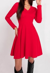 Rotes Langarmkleid mit V-Ausschnitt, tailliertem Oberteil, weitem Rock und glatter Textur, kombiniert mit knielangen schwarzen Stiefeln.