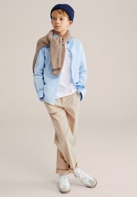 Šviesiai mėlyna palaidinė, balti marškinėliai, smėlio spalvos kelnės, ant pečių permesta rusvos spalvos megzta palaidinė, tamsiai mėlynas megztas kepurės bžūp bei balti sportbačiai.