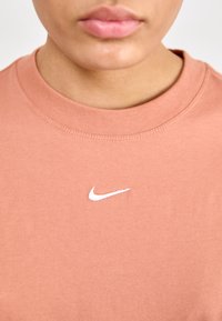 Růžové bavlněné tričko s kulatým výstřihem a bílým logem Nike umístěným ve středu, s hladkou texturou a standardním střihem.