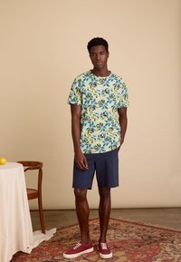 Lichtblauw T-shirt met korte mouwen met een bloemenpatroon in geel en groen, gecombineerd met marineblauwe shorts en bordeauxrode sneakers.