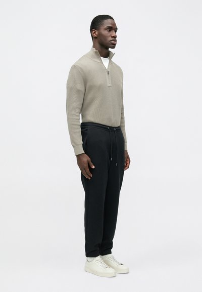 Ανοιχτό μπεζ, ριμπ πλεκτό πουλόβερ με φερμουάρ, συνδυασμένο με μαύρο παντελόνι φόρμας με κορδόνι. Το μοντέλο φοράει άσπρα αθλητικά παπούτσια. Απλός, μοντέρνος σχεδιασμός.