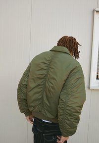 Giacca bomber verde kaki con zip frontale, polsini a costine e cuciture arricciate lungo la schiena, realizzata in un tessuto lucido e texture.