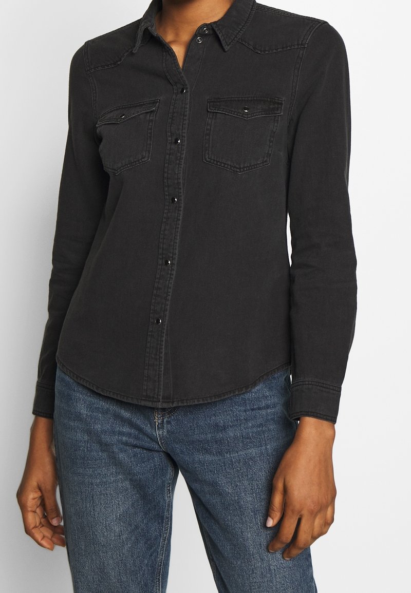 Personne portant une chemise noire à boutons avec des poches poitrine et un jean en denim bleu, debout devant un fond blanc uni.