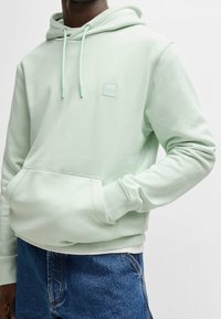 Mátově zelená mikina s kapucí z měkké látky, vybavená přední kapsou, stahovací šňůrkou a malým vyraženým logem na hrudi.