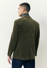 Hombre vestido con un blazer de pana verde oliva y jeans azul oscuros, mirando hacia la izquierda frente a un fondo claro.
