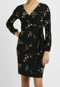 Svart omlottklänning med långa ärmar, V-halsringning och ett blommönster av vita och gula blommor. Mjuk tyg med en slät yta.
